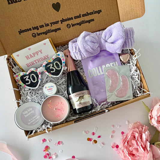 A curated pamper 30th gift box  includes a Collagen face mask sheet by Lapcos, purple under eye masks, , a pink birthday candle, a pink chocolate heart,  a small bottle of sparkling rosé, , a purple headband , birthday cake lipbalm and a happy birthday chocolate bar.