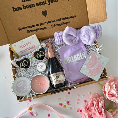 A curated birthday gift box filled with spa and self-care items, photographed from above on a white surface with pink flowers around it. The open kraft box lid has printed text and social handles. Prosecco , happy birthday chocoalte bar, 40th glasses, candle, lapcos facemask, under eye, towelling headband, birthday cake lipbalm, chocolate heart.