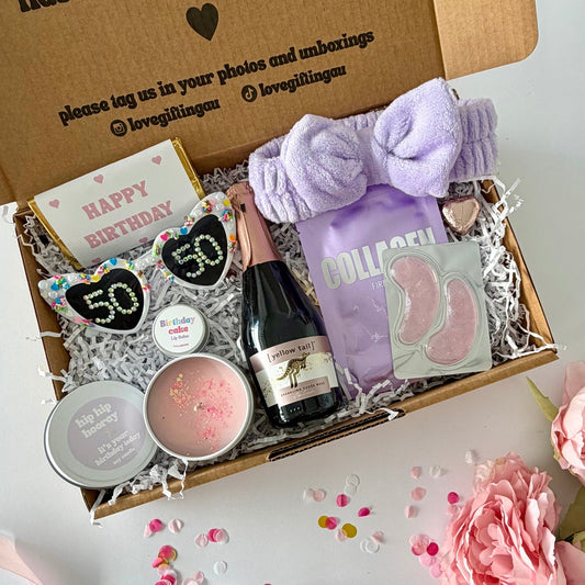 A curated birthday 50th gift box filled with spa and self-care items, photographed from above on a white surface with pink flowers around it. The open kraft box lid has printed text and social handles. Prosecco , happy birthday chocoalte bar, 50th glasses, candle, lapcos facemask, under eye, towelling headband, birthday cake lipbalm, chocolate heart.