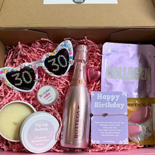 Open box containing a bottle of champagne, sunglasses with '30' on them, and other birthday-themed items.