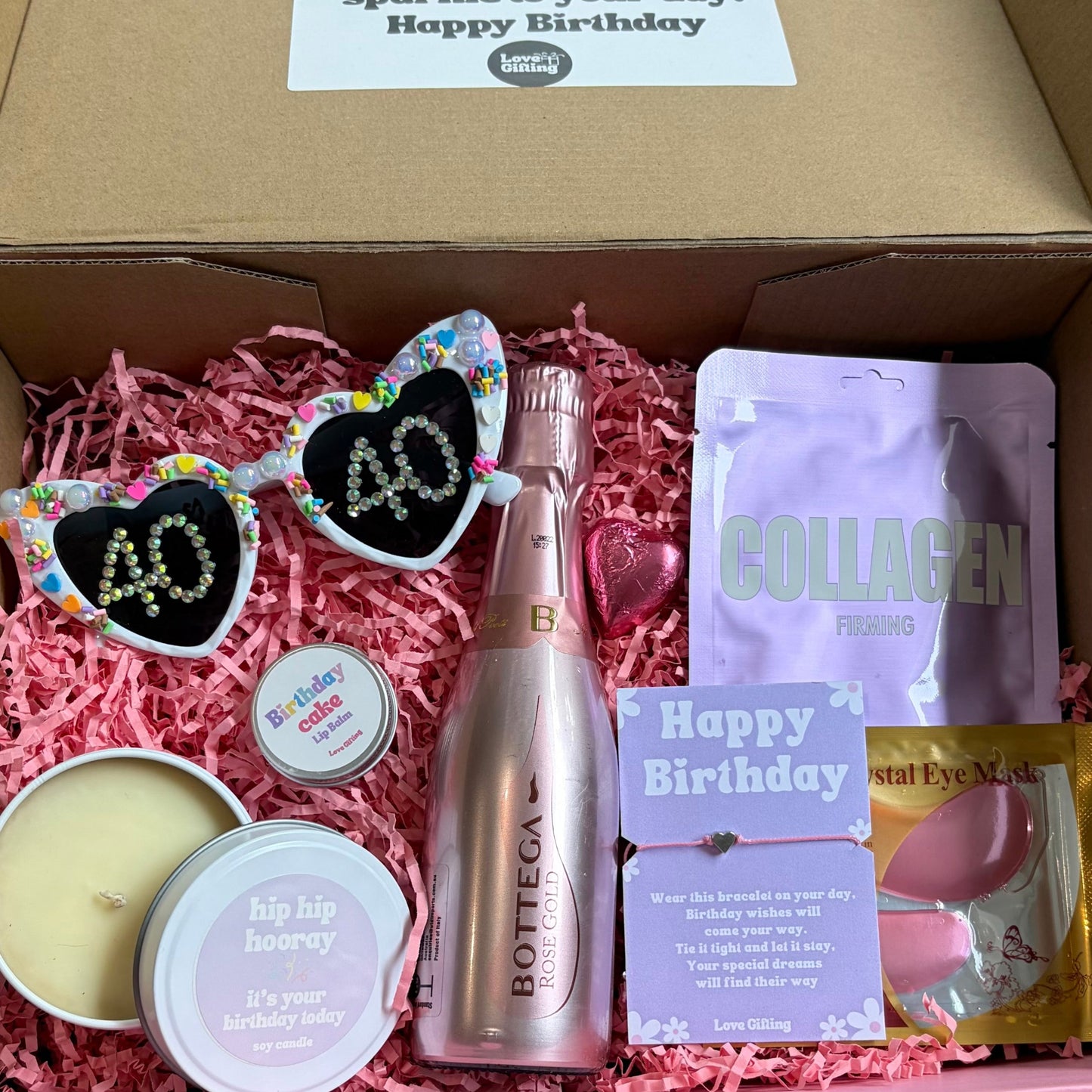Open box containing birthday-themed items including a bottle, sunglasses, and packets on a wooden surface.