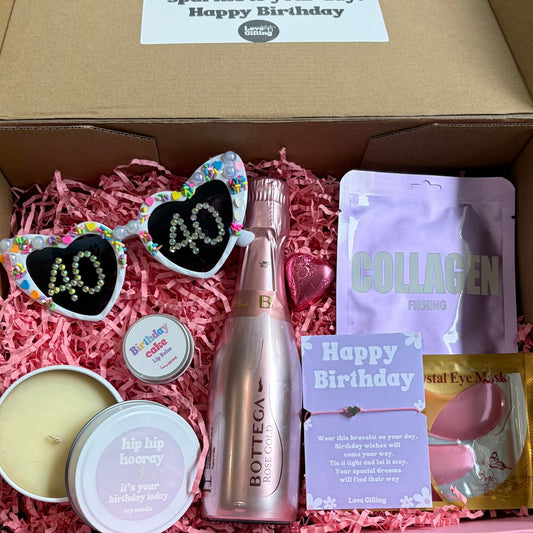 Open box containing birthday-themed items including a bottle, sunglasses, and packets on a wooden surface.