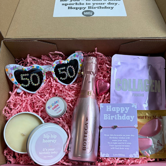 Gift box with birthday-themed items including a bottle, sunglasses, and snacks on a wooden surface.