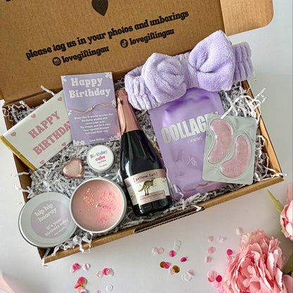 A curated birthday gift box filled with spa and self-care items, photographed from above on a white surface with pink flowers around it. The open kraft box lid has printed text and social handles. Prosecco , happy birthday chocoalte bar, candle, lapcos facemask, under eye, towelling headband, birthday cake lipbalm, chocolate heart. Choice of birthday sunglasses or happy birtnday bracelet