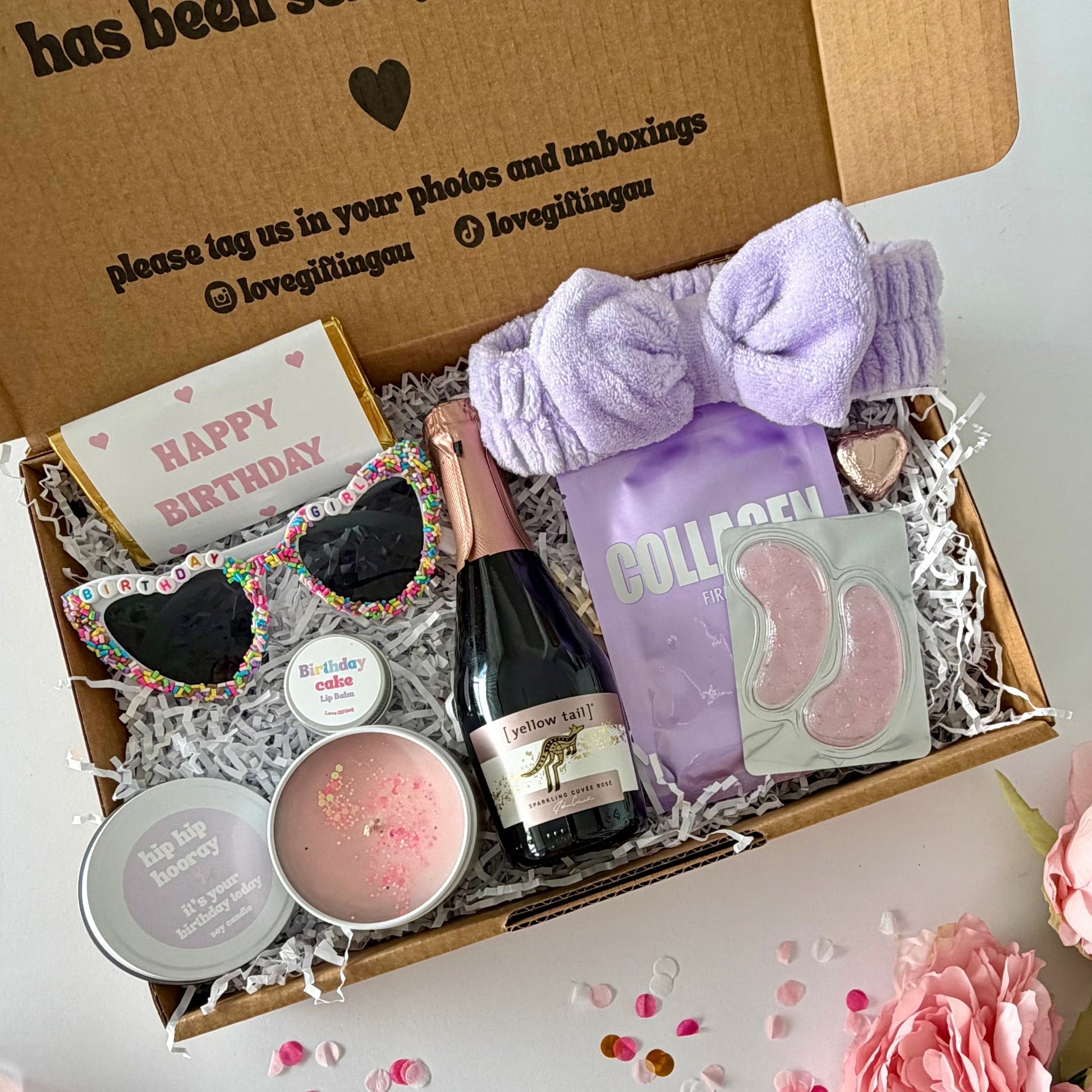 A curated birthday gift box filled with spa and self-care items, photographed from above on a white surface with pink flowers around it. The open kraft box lid has printed text and social handles. Prosecco , happy birthday chocoalte bar, birthday glasses, candle, lapcos facemask, under eye, towelling headband, birthday cake lipbalm, chocolate heart.