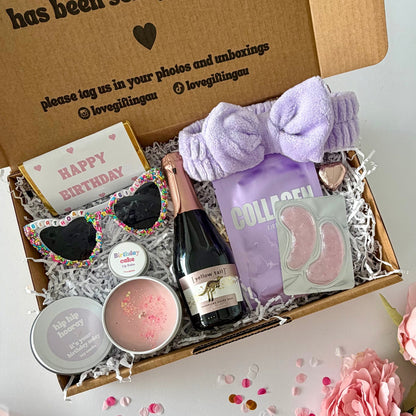 A curated birthday gift box filled with spa and self-care items, photographed from above on a white surface with pink flowers around it. The open kraft box lid has printed text and social handles. Prosecco , happy birthday chocoalte bar, birthday glasses, candle, lapcos facemask, under eye, towelling headband, birthday cake lipbalm, chocolate heart.