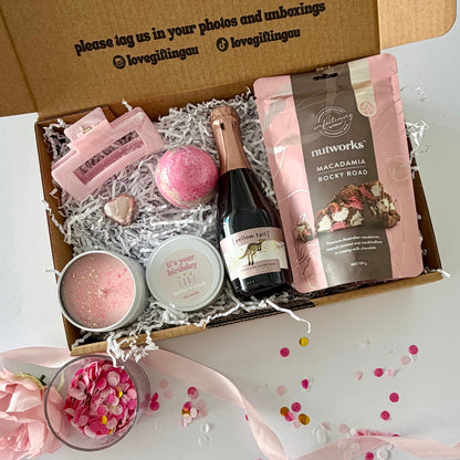 A curated pamper  gift box  includes a starwberry bath bomb, a pink candle, a pink chocolate heart,  a small bottle of sparkling rosé, , a bag of Nutworks macadamia rocky road, and a pink claw hair clip