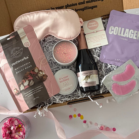 A curated gift box set displayed in an open brown cardboard box with printed message text on the lid. Inside are a pink satin sleep mask, a pink candle tin, a bag of Nutworks rocky road, lapcos collagen sheet mask, and pink eye patches, birthday bracelet arranged on white filler with pink ribbon and flower petals around it.