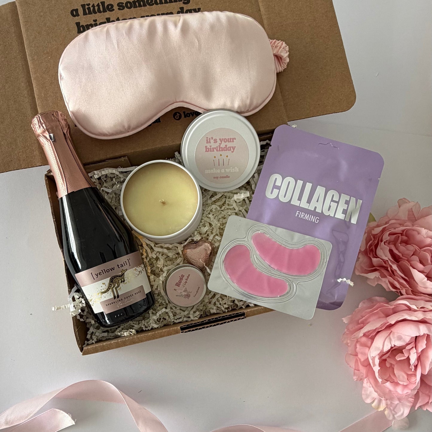 A curated gift box set on a white surface, featuring a light pink silk sleep mask, a bottle of pink champagne, a pink candle, a sheet of pink eye masks, and a purple collagen supplement pouch, arranged with a pink ribbon.