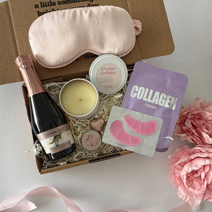 A curated gift box set on a white surface, featuring a light pink silk sleep mask, a bottle of pink champagne, a pink candle, a sheet of pink eye masks, and a purple collagen supplement pouch, arranged with a pink ribbon.