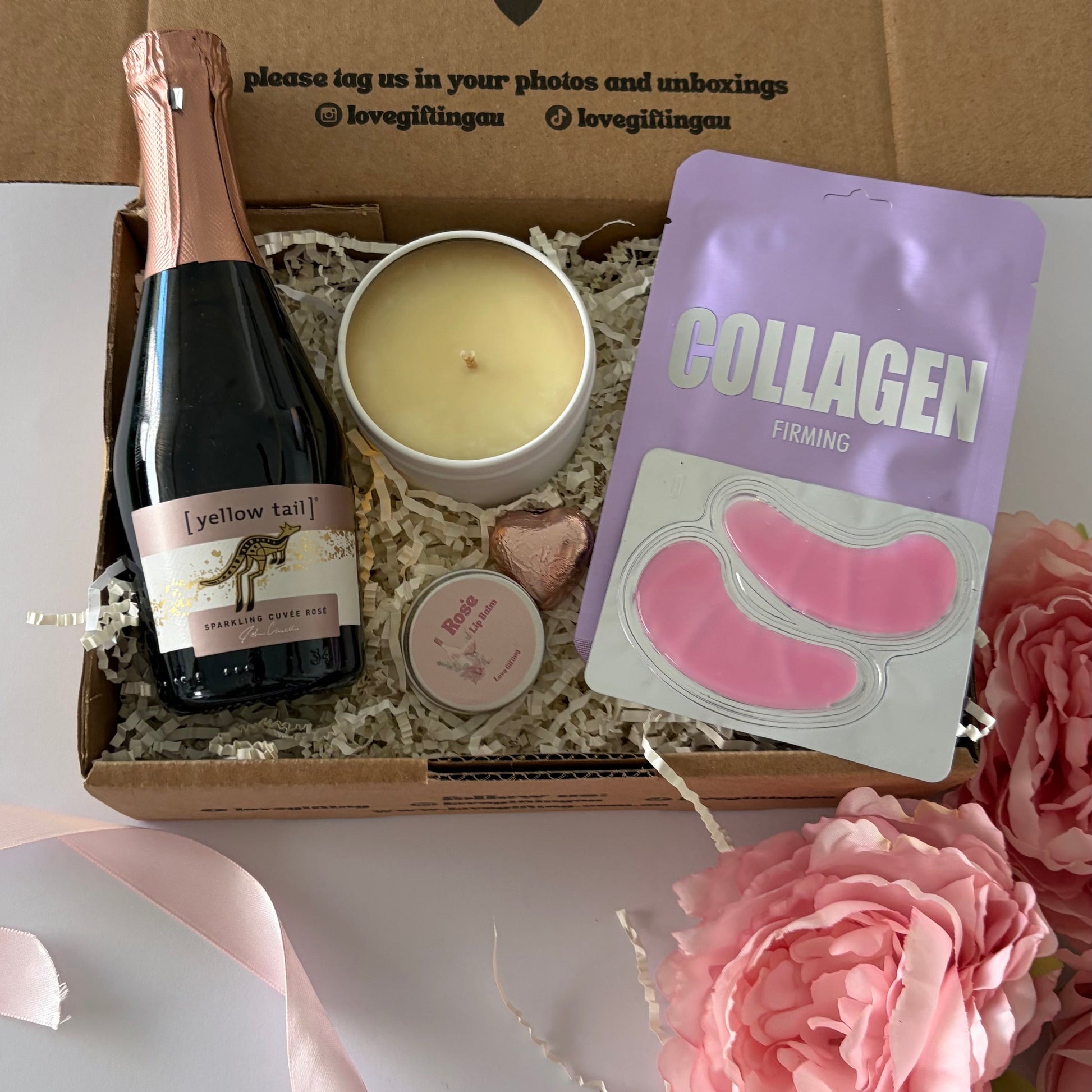 A curated gift box set displayed in an open brown cardboard box with printed message inside. The set includes a bottle of champagne, a candle, a pink heart-shaped item, a pink eye mask packet labeled 'COLLAGEN', and a small round tin.