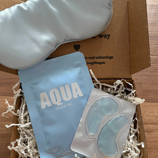 Box containing a white eye mask, blue pouch labeled 'Aqua', and clear gel pads on a wooden surface.