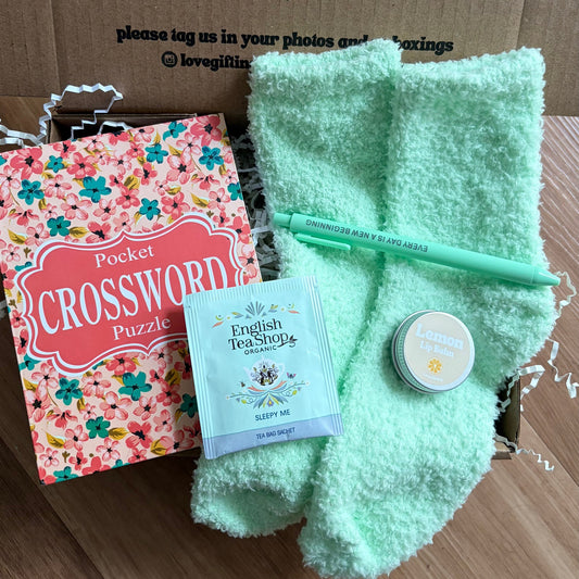Gift box with green socks, tea, crossword puzzle, and candle on a wooden surface.