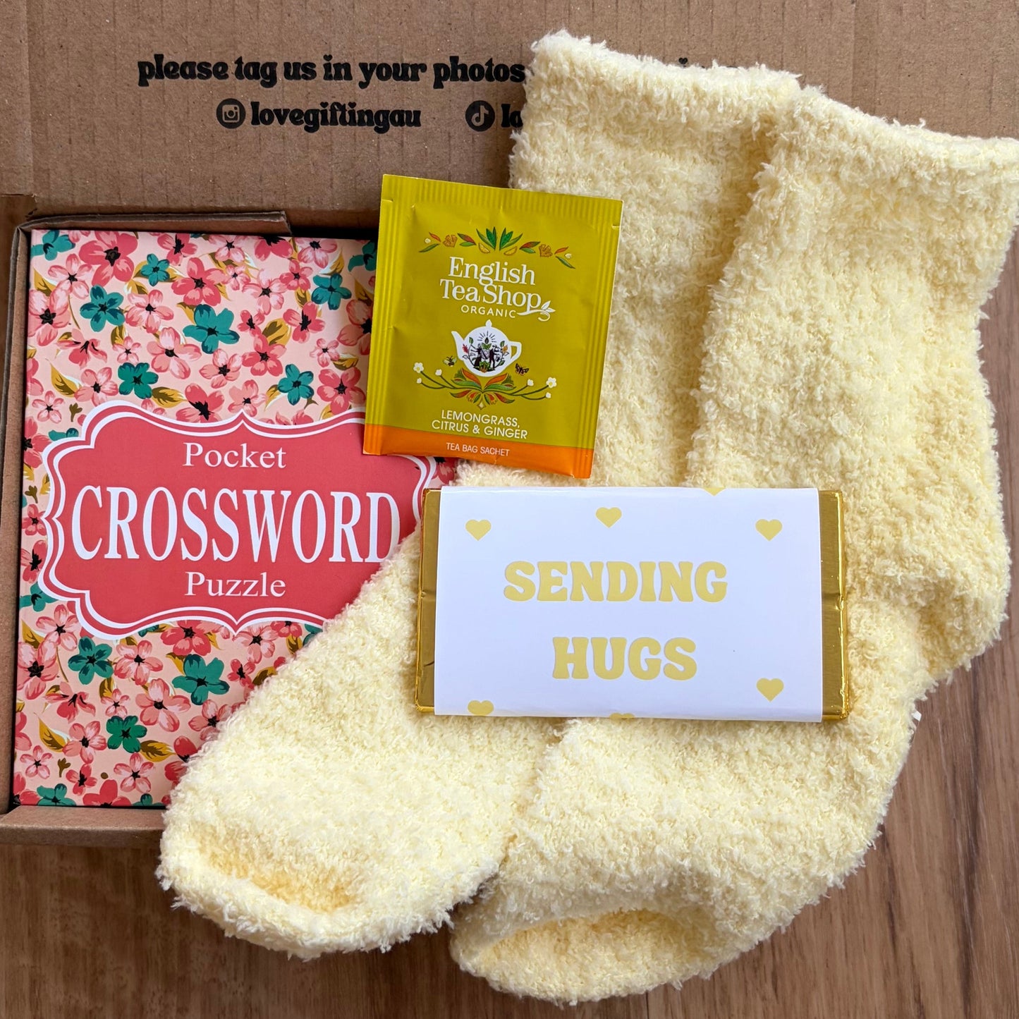 Box of tea, crossword puzzle, and card with 'Sending Hugs' message on a cardboard background.