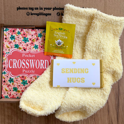 Box of tea, crossword puzzle, and card with 'Sending Hugs' message on a cardboard background.