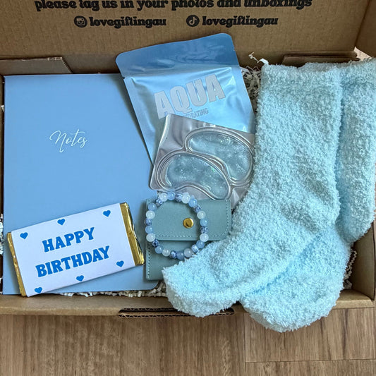 Box of birthday-themed items including socks, a card, and a chocolate bar on a wooden surface.