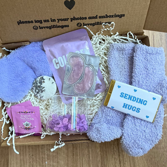 Box of care package with plush toys, cards, and a small plant on a wooden floor.