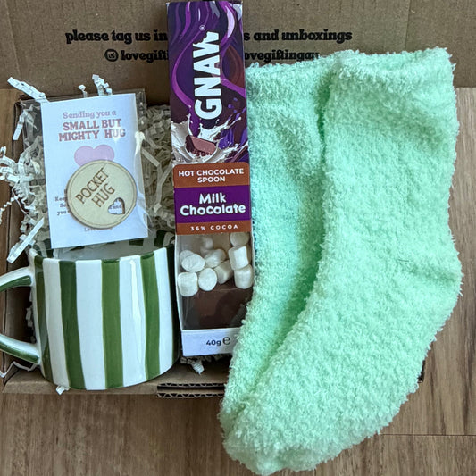 Box of care package with a green sock, mug, and chocolate bar on a wooden surface.