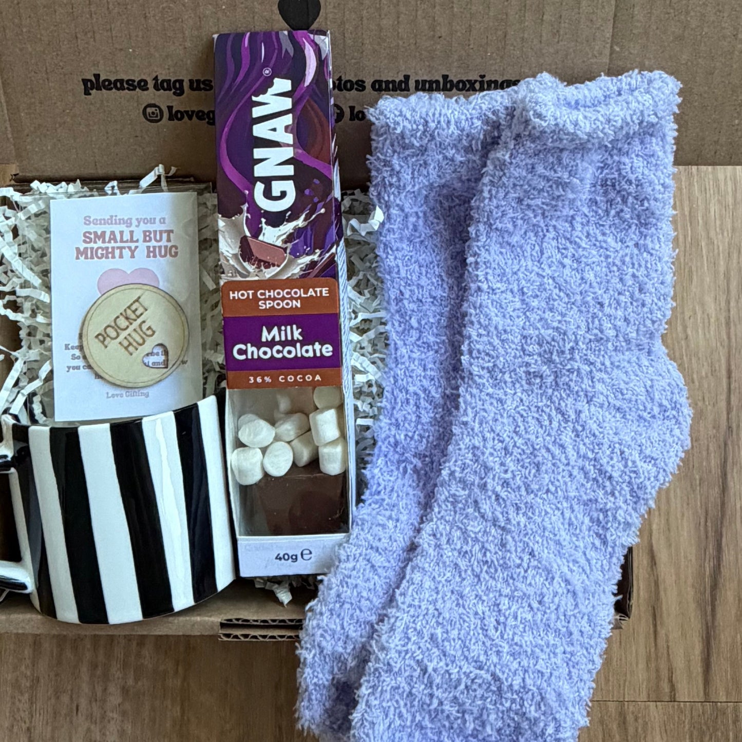 Box of assorted items including socks, chocolate, and a mug on a wooden surface.