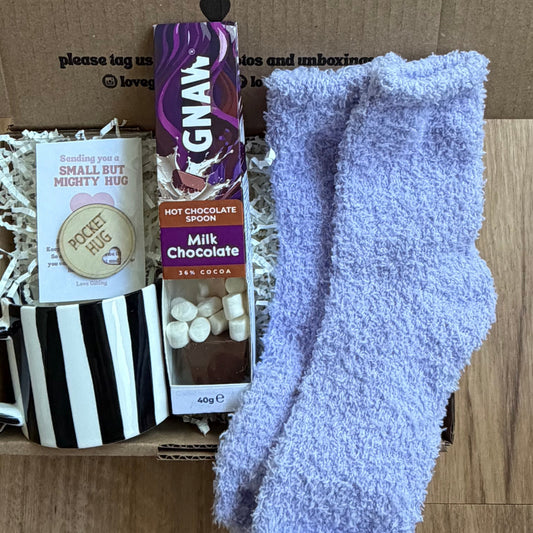 Box of assorted items including socks, chocolate, and a mug on a wooden surface.