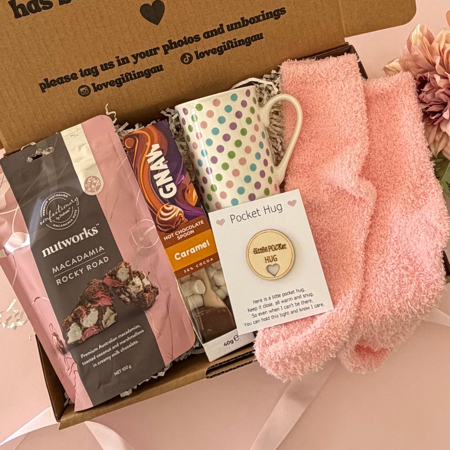 A curated gift box set containing a pink fleece pocket hug, a patterned ceramic mug, a bag of Nutworks chocolate, a bar of Ganlon chocolate, and a printed message card inside a brown cardboard box.