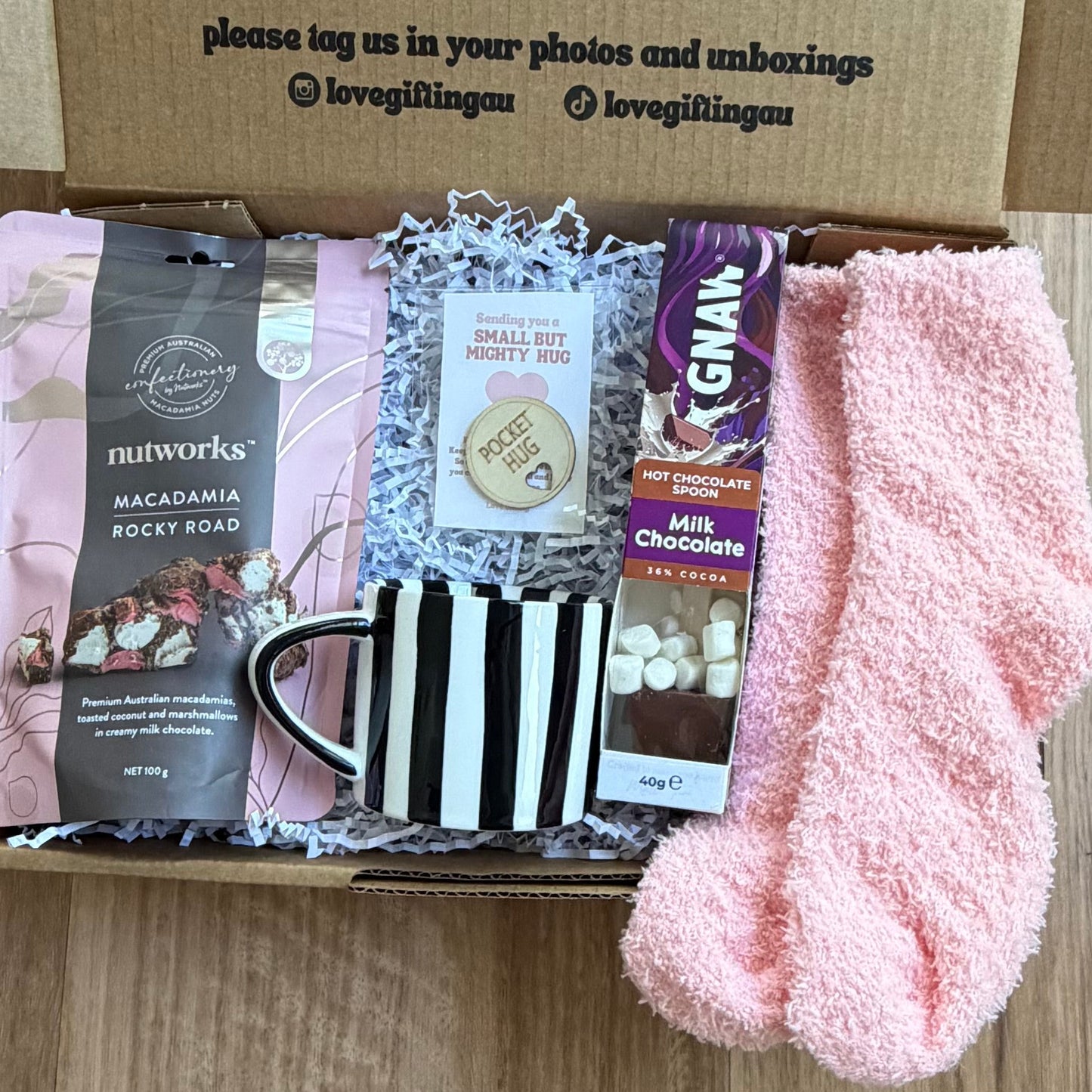 Box of care items including a mug, pink socks, and other small packages on a wooden surface.