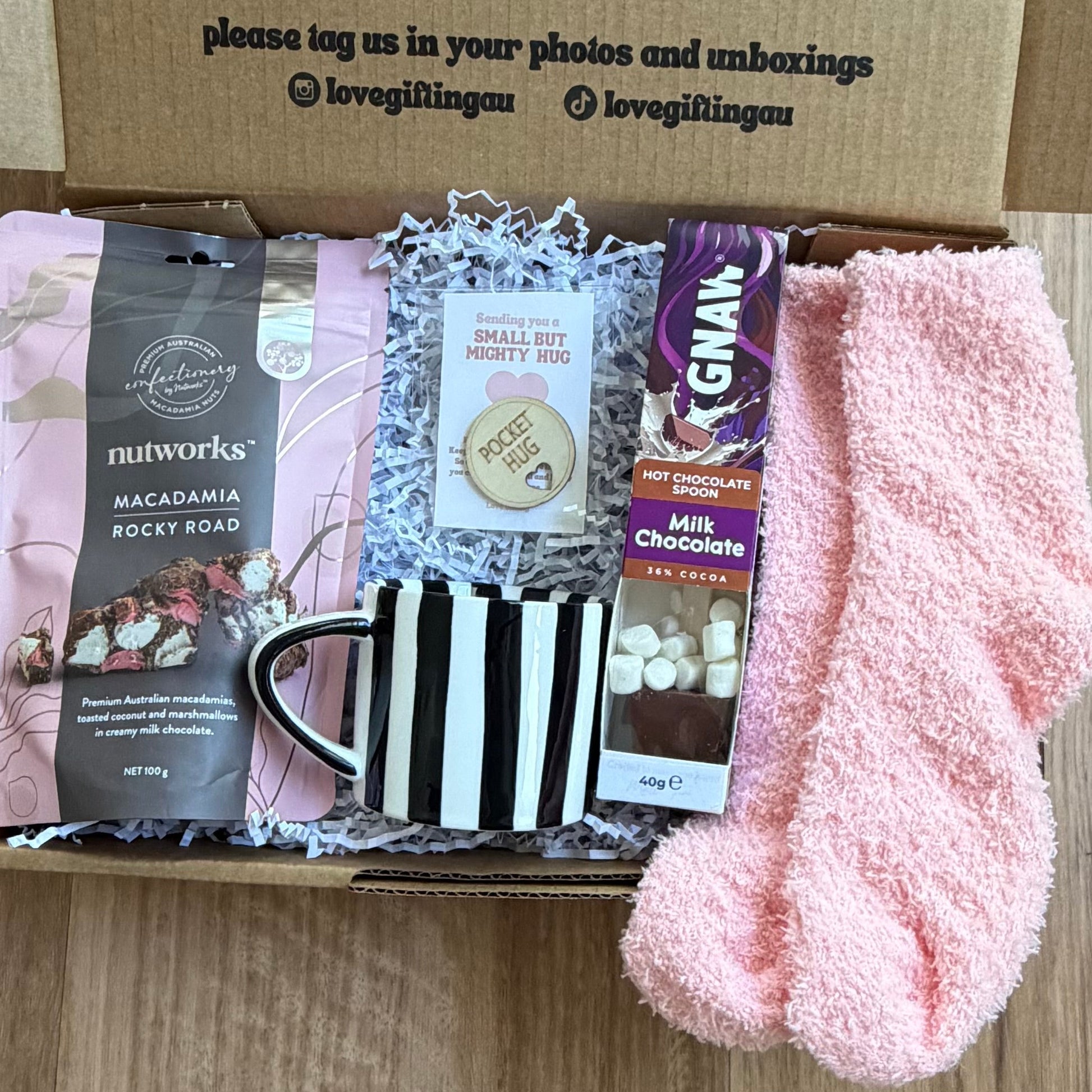 Box of care items including a mug, pink socks, and other small packages on a wooden surface.