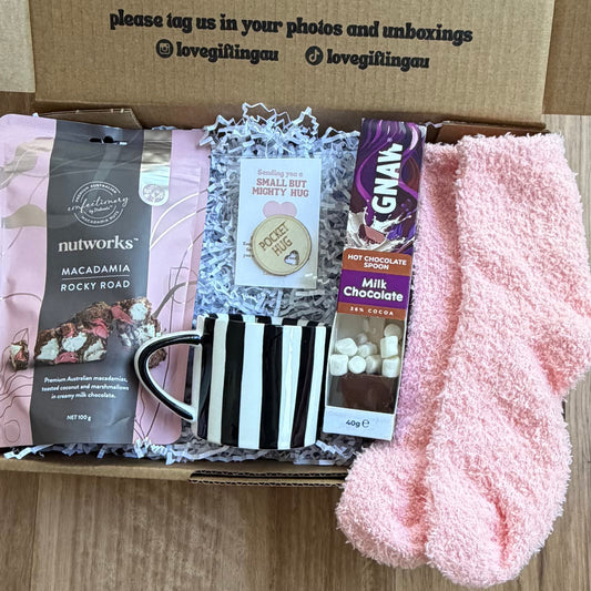 Box of care items including a mug, pink socks, and other small packages on a wooden surface.