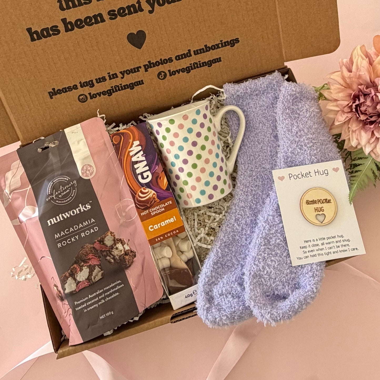 A curated gift box set on a pale pink background, featuring a brown cardboard box printed with a message, a pink and white bag of Morwark's chocolate, a white mug with green dots, a lavender pocket hug sock, and a pink flower.