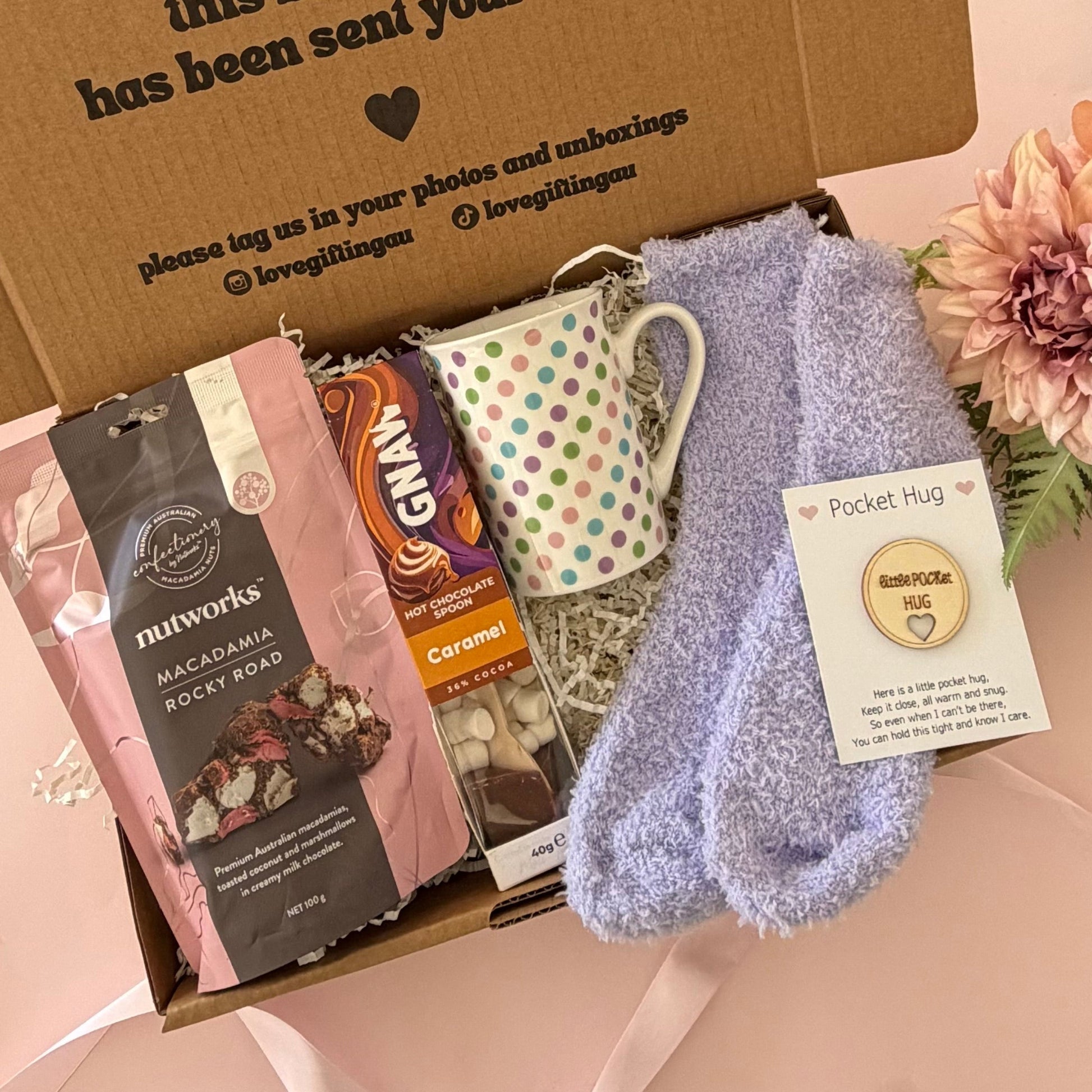 A curated gift box set on a pale pink background, featuring a brown cardboard box printed with a message, a pink and white bag of Morwark's chocolate, a white mug with green dots, a lavender pocket hug sock, and a pink flower.