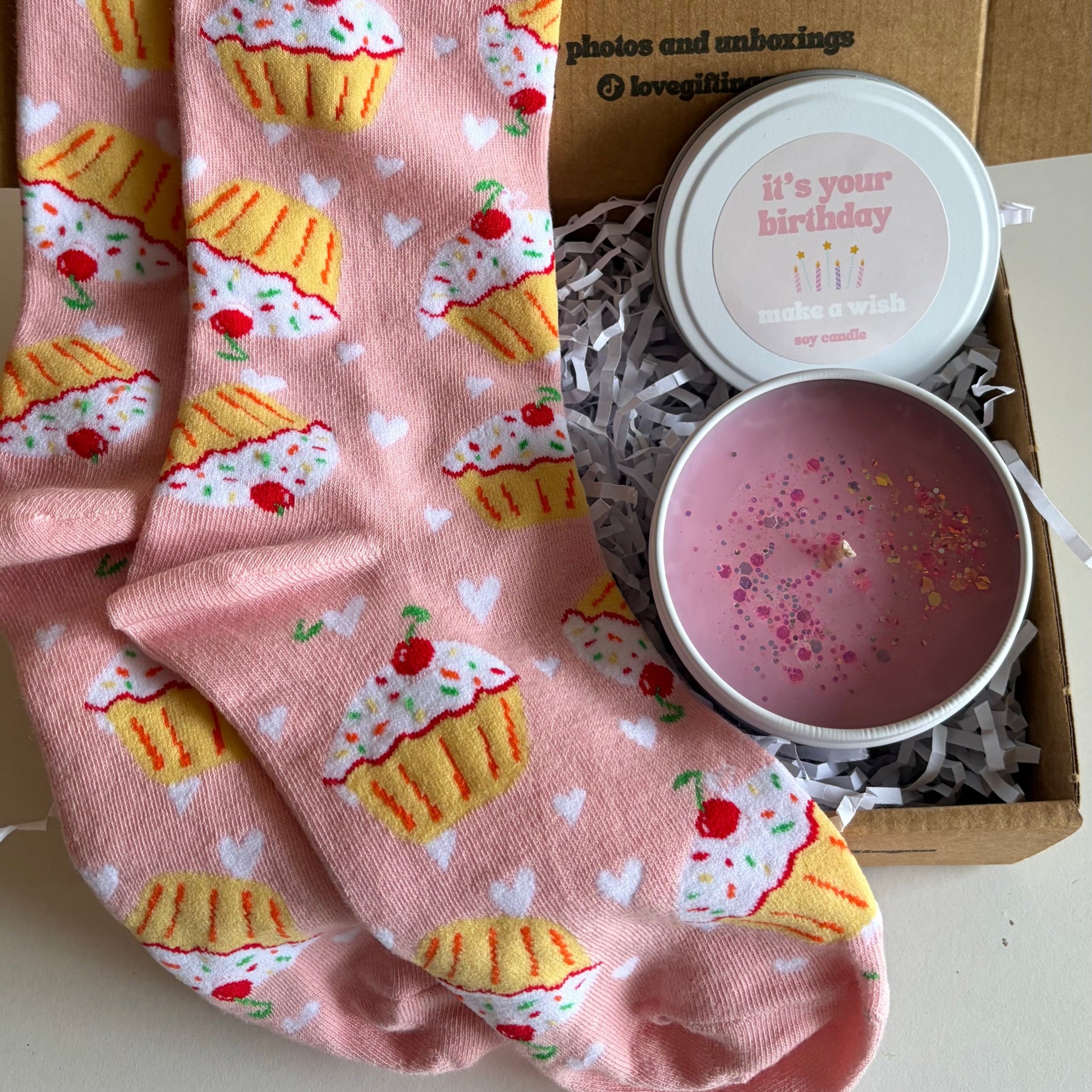Pink socks with cupcake design and a pink candle in a box on a white surface.