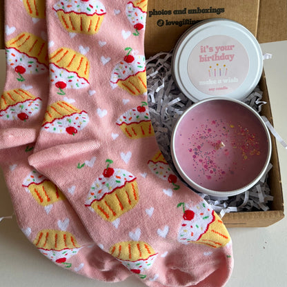 Pink socks with cupcake design and a pink candle in a box on a white surface.