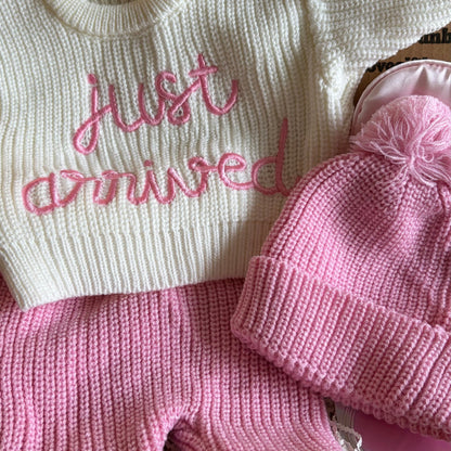 Baby outfit with 'just arrived' text, pink hat, and pants on a cardboard box background.