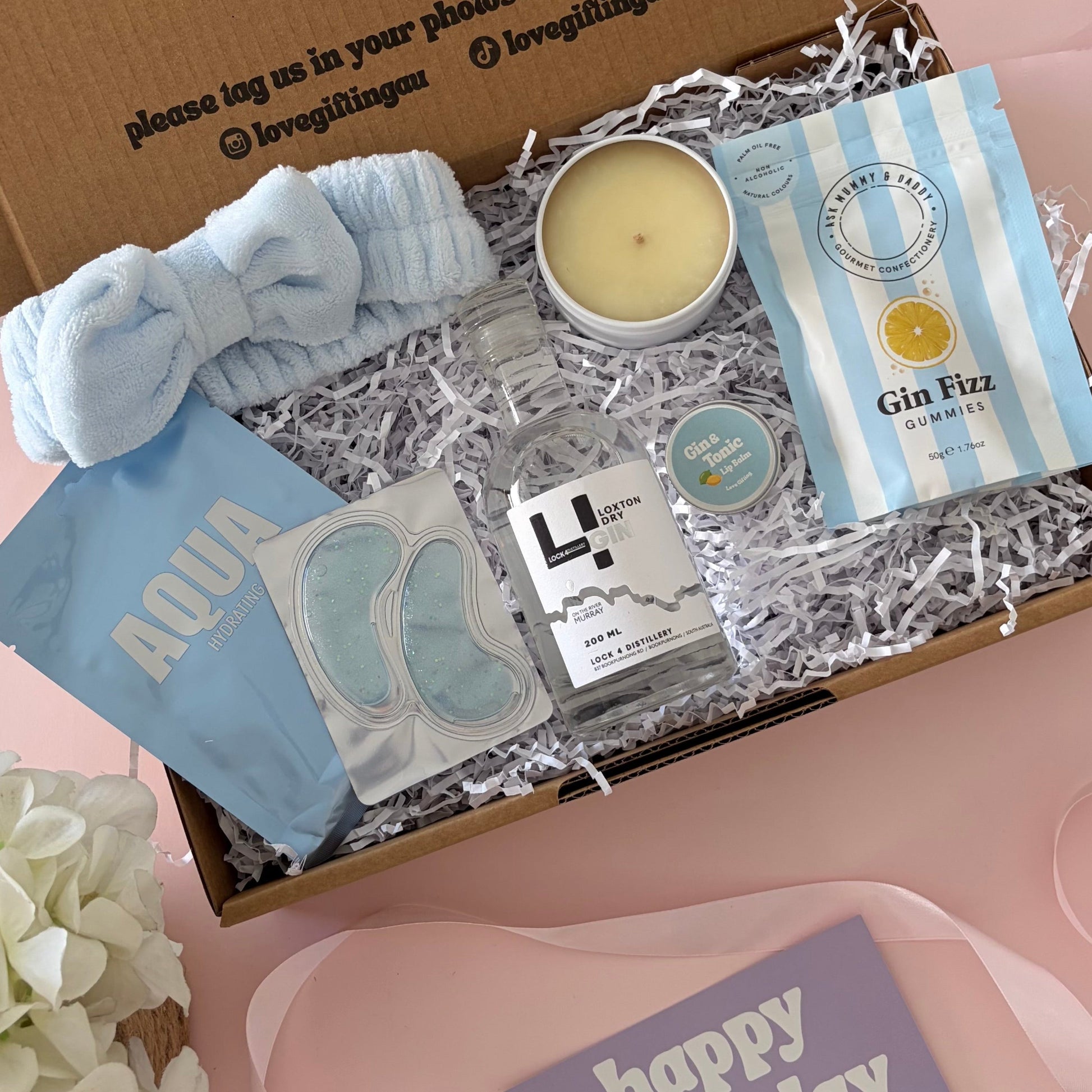A curated gift box set displayed on a pink surface, featuring a brown cardboard box with printed text, a light blue scrunchie, a candle, packaged bath items, and a purple 'happy birthday' card. A white flower arrangement sits at the lower left.
