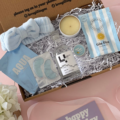 A curated gift box set displayed on a pink surface, featuring a brown cardboard box with printed text, a light blue scrunchie, a candle, packaged bath items, and a purple 'happy birthday' card. A white flower arrangement sits at the lower left.
