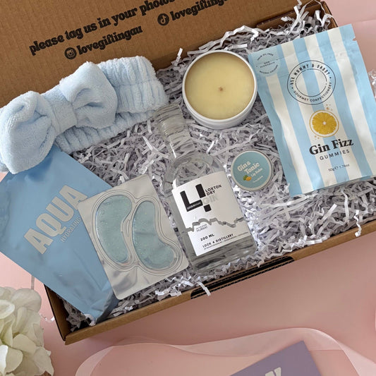 A curated gift box set displayed on a pink surface, featuring a brown cardboard box with printed text, a light blue scrunchie, a candle, packaged bath items, and a purple 'happy birthday' card. A white flower arrangement sits at the lower left.