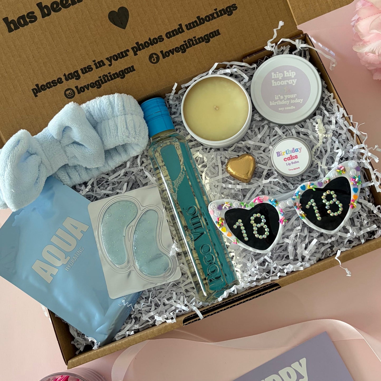 A curated 18th birthday gift box . The box contains a light blue baby headband a mini  bottle of sav blanc, a small candle, lapcos face mask , under eye mask ,lipbalm and age '18'  sunglasses 