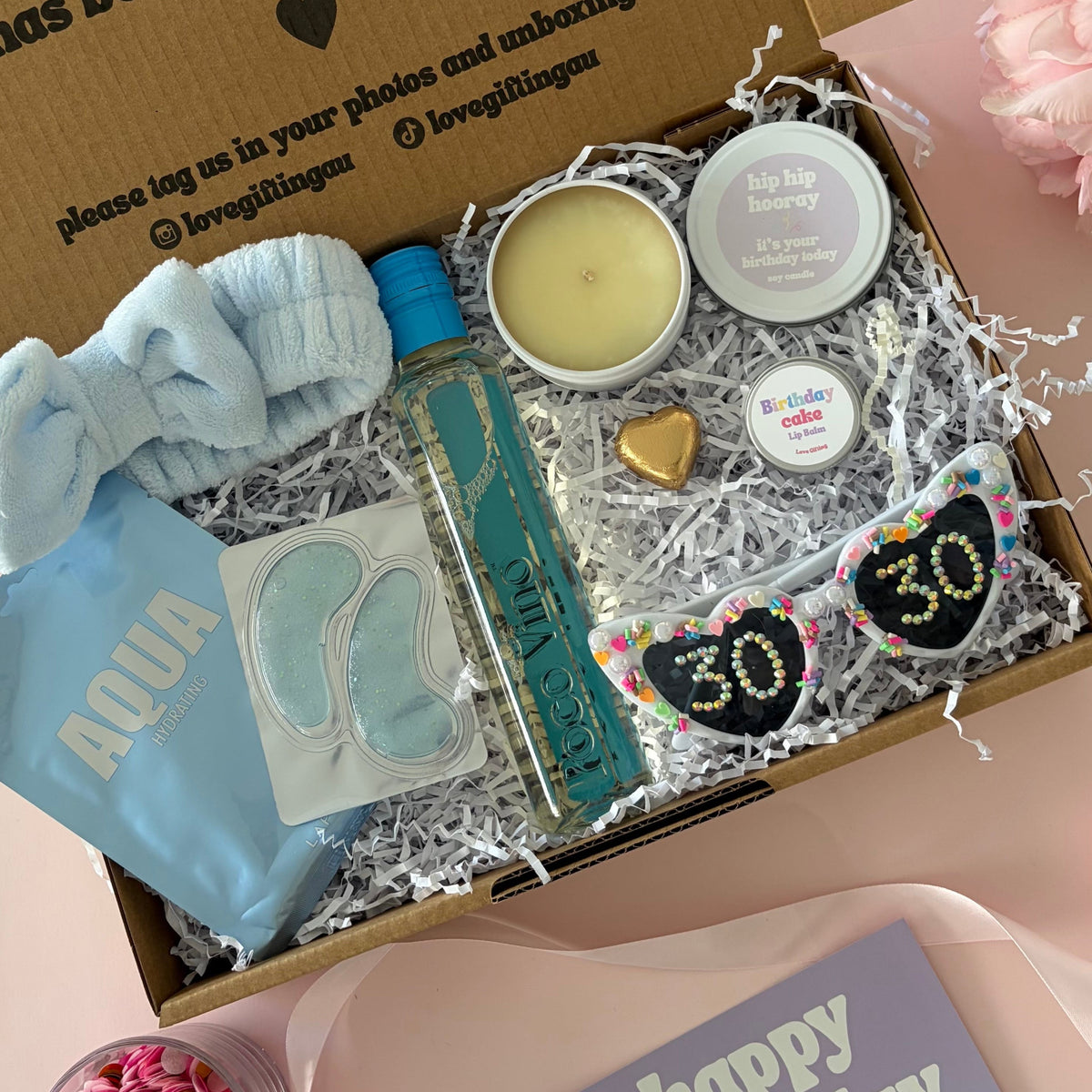 A curated 30th birthday gift box . The box contains a light blue baby headband a mini  bottle of sav blanc, a small candle, lapcos face mask , under eye mask ,lipbalm and age '30'  sunglasses 