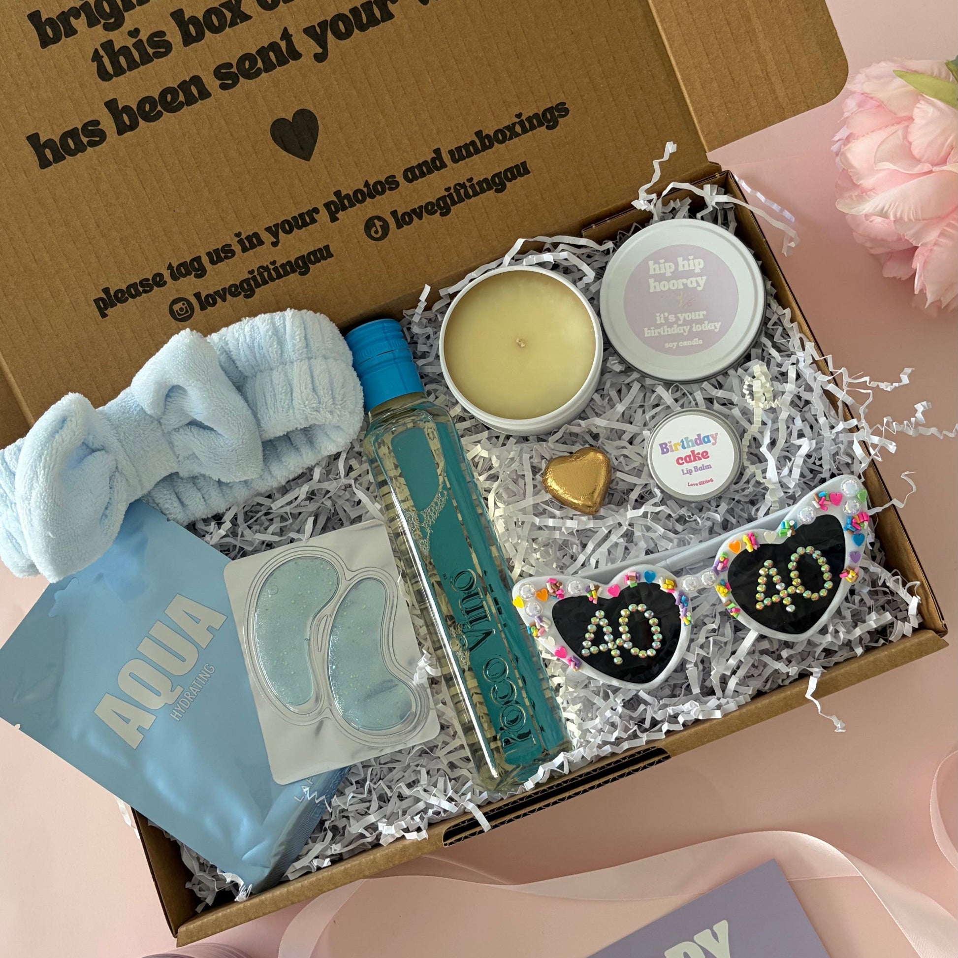 A curated 40th birthday gift box . The box contains a light blue baby headband a mini  bottle of sav blanc, a small candle, lapcos face mask , under eye mask ,lipbalm and age '30'  sunglasses 