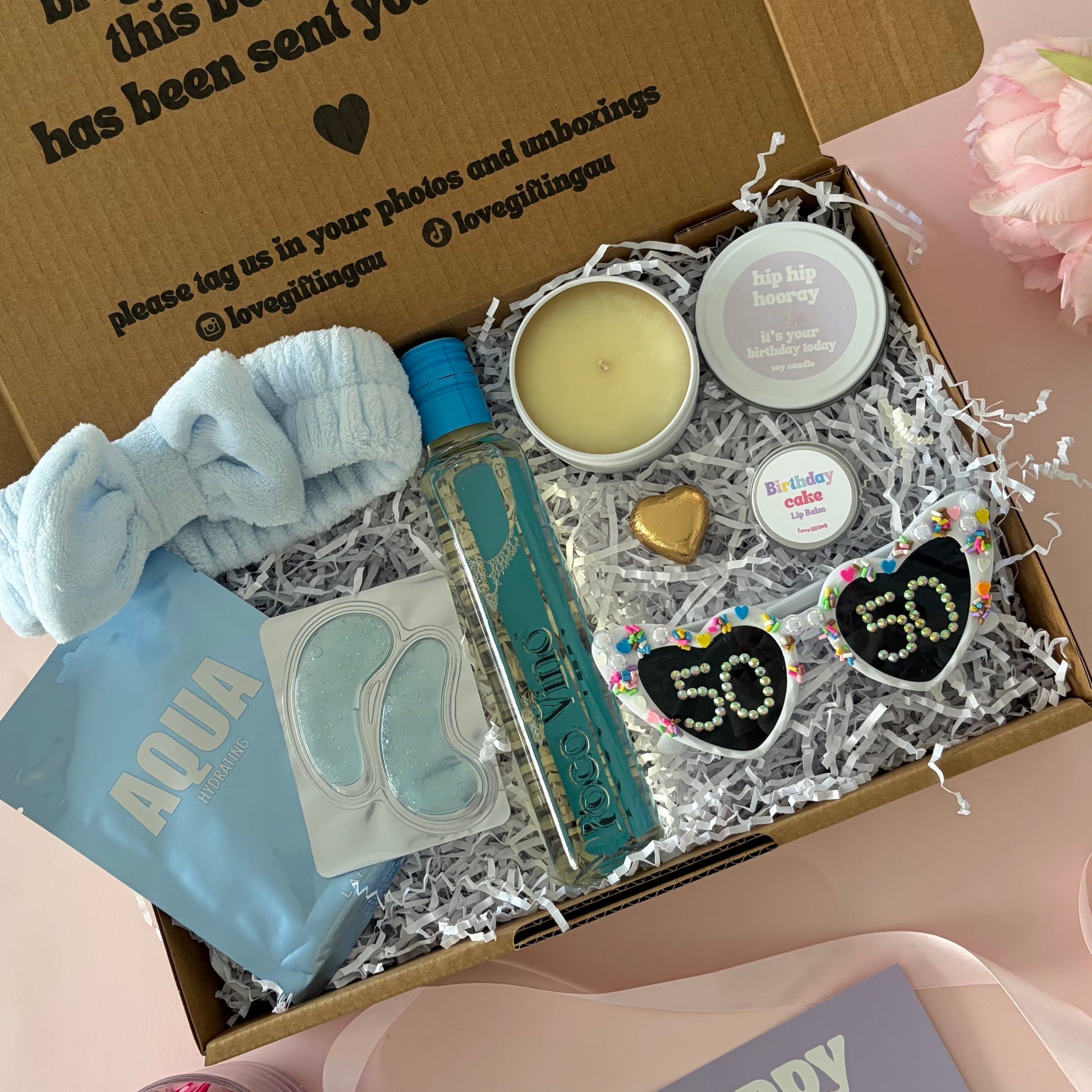 A curated 50th birthday gift box . The box contains a light blue baby headband a mini  bottle of sav blanc, a small candle, lapcos face mask , under eye mask ,lipbalm and age '50'  sunglasses 