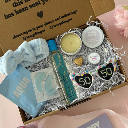 A curated 50th birthday gift box . The box contains a light blue baby headband a mini  bottle of sav blanc, a small candle, lapcos face mask , under eye mask ,lipbalm and age '50'  sunglasses 