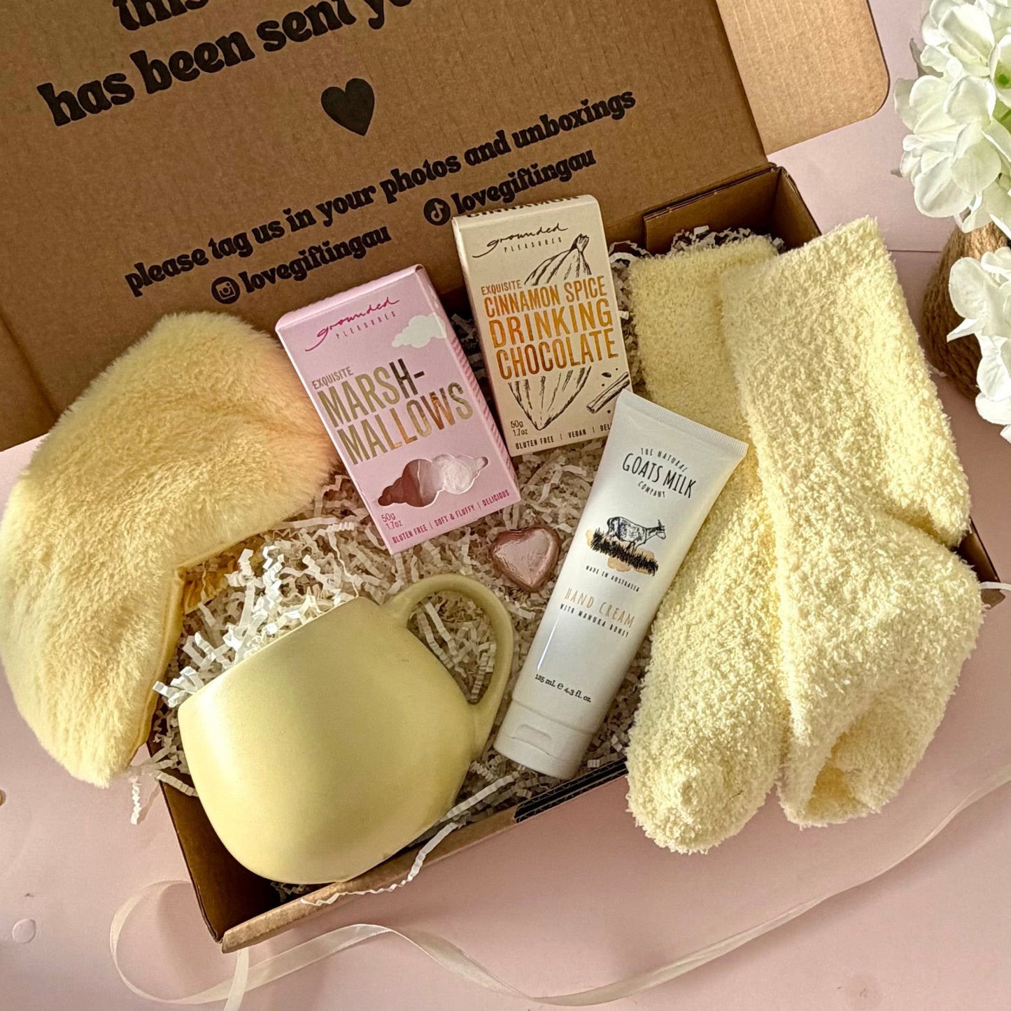 A curated gift box featuring a pale yellow mug, fluffy yellow socks, pink marshmallow box a  cinamon hot chocolate marshmallows, mini chocolate heart, fluffy sleep mask and a  tube of hand cream  
