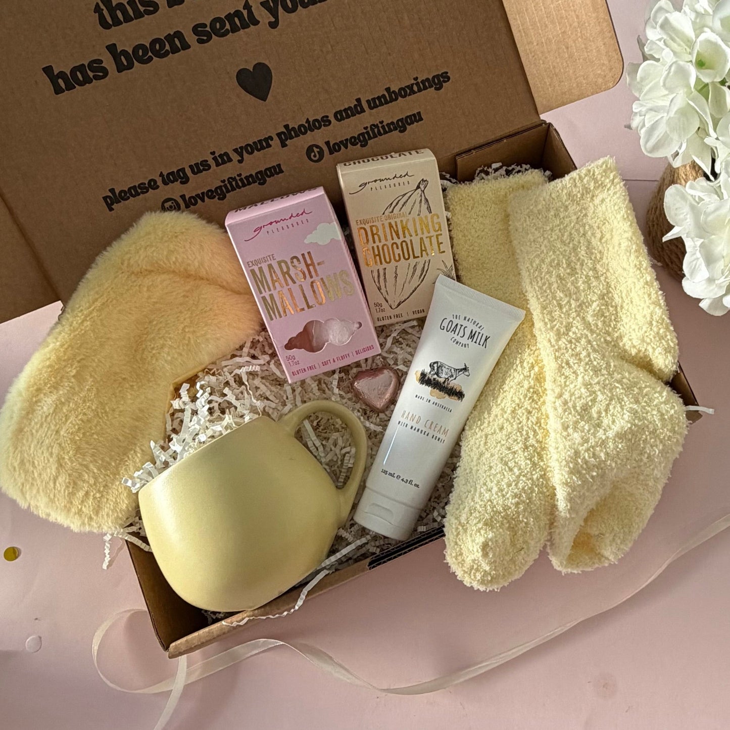A curated gift box featuring a pale yellow mug, fluffy yellow socks, pink marshmallow box a  hot chocolate marshmallows, mini chocolate heart, fluffy sleep mask and a  tube of hand cream  