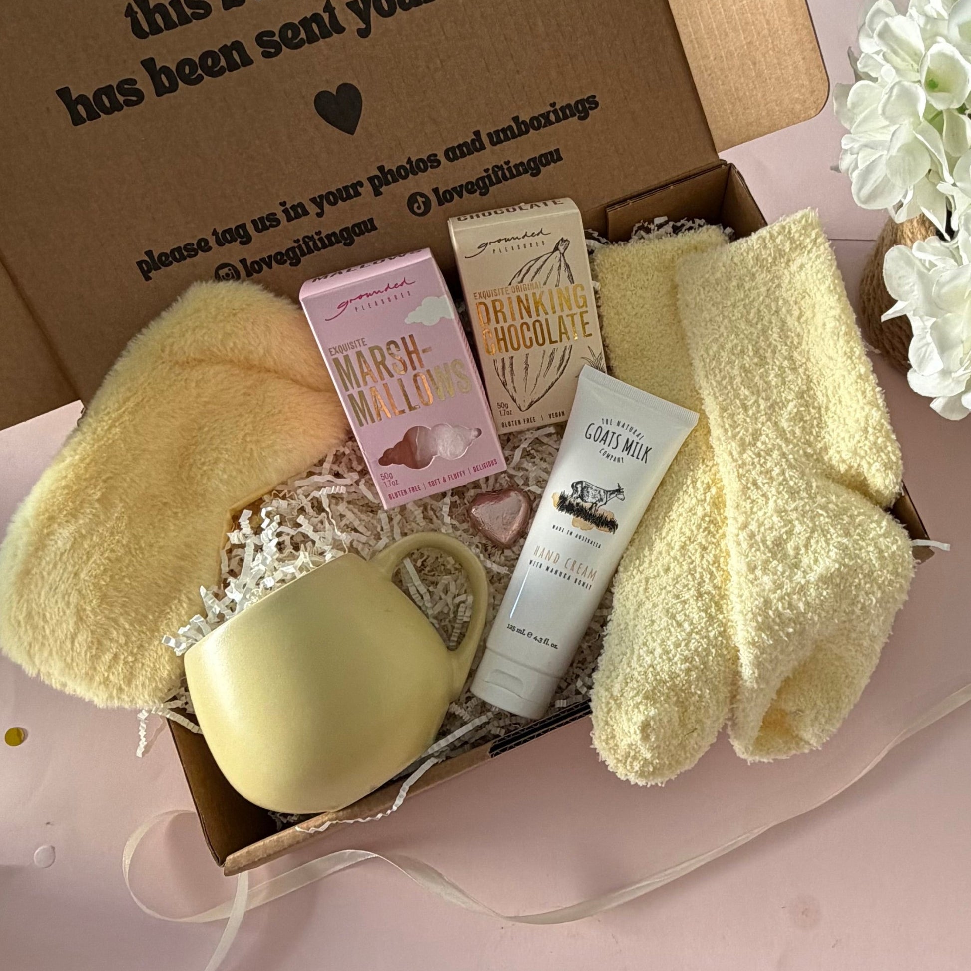 A curated gift box featuring a pale yellow mug, fluffy yellow socks, pink marshmallow box a  hot chocolate marshmallows, mini chocolate heart, fluffy sleep mask and a  tube of hand cream  