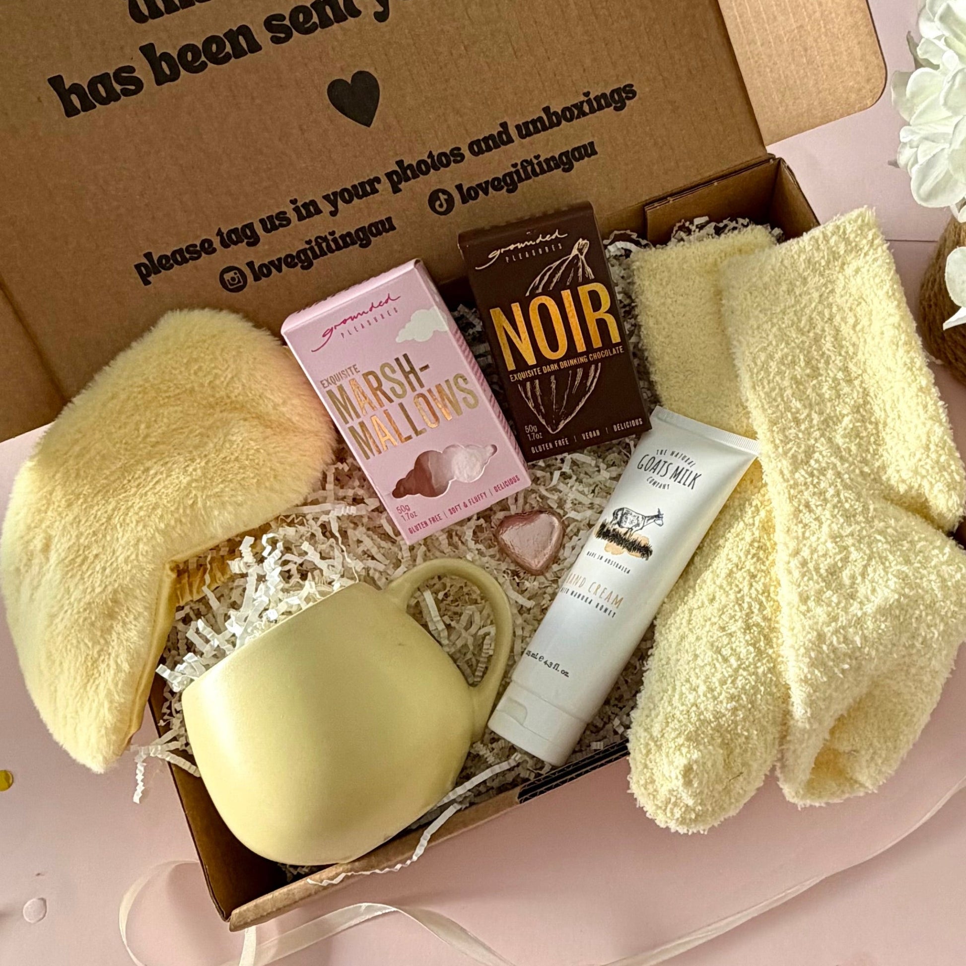 A curated gift box featuring a pale yellow mug, fluffy yellow socks, pink marshmallow box a dark hot chocolate labeled 'NOIR', marshmallows, mini chocolate heart, fluffy sleep mask and a  tube of hand cream  