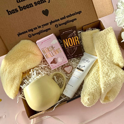 A curated gift box featuring a pale yellow mug, fluffy yellow socks, pink marshmallow box a dark hot chocolate labeled 'NOIR', marshmallows, mini chocolate heart, fluffy sleep mask and a  tube of hand cream  