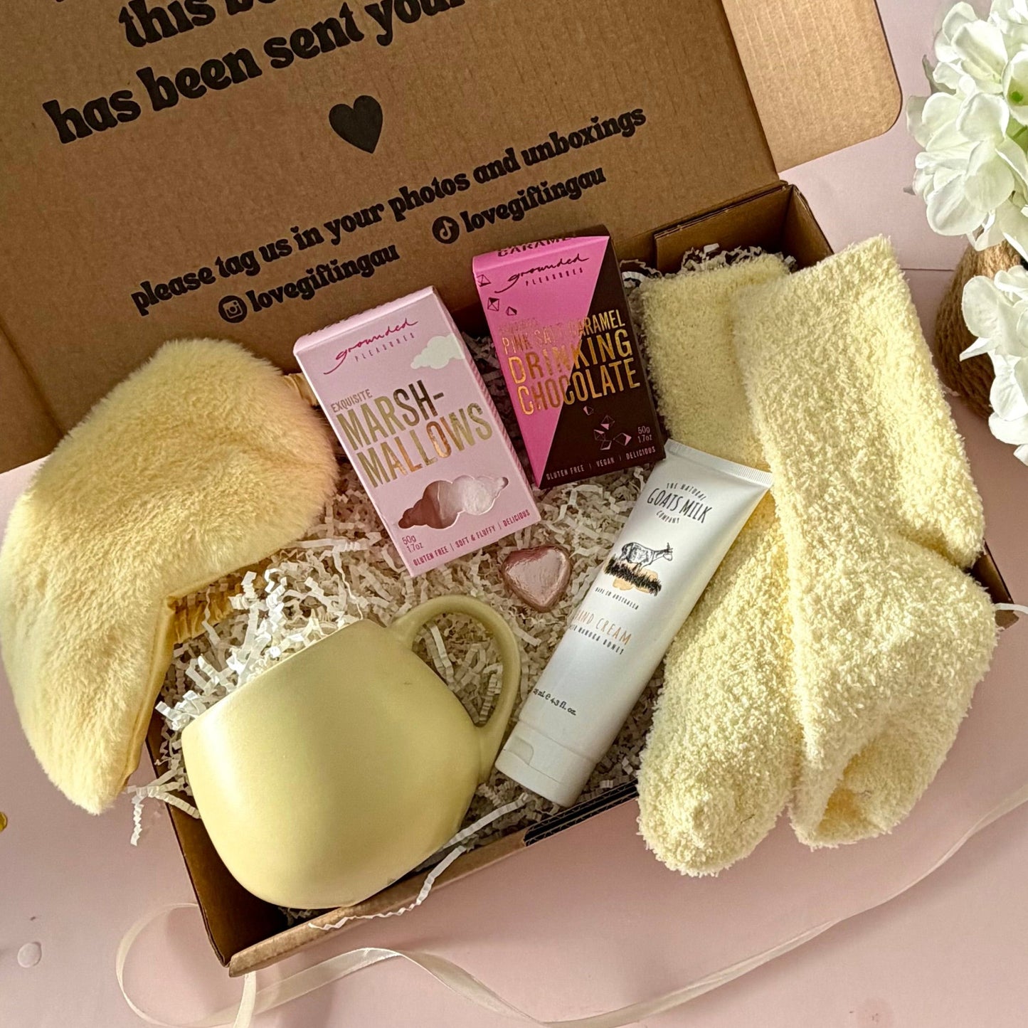 A curated gift box featuring a pale yellow mug, fluffy yellow socks, grounded pleasures pink marshmallow box a pink salt and caramel hot chocolate marshmallows, mini chocolate heart, fluffy sleep mask and a  tube of hand cream  