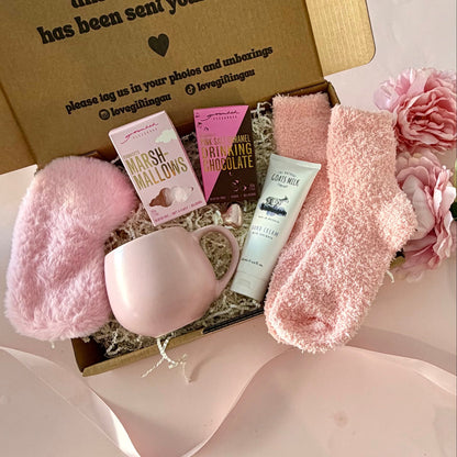 A curated comfort winter gift box featuring a pink mug, fluffy pink socks, gourmet marshmallow box, Grounded Pleasures hot chocolate box, mini chocolate heart, pink fluffy sleep mask and a lavender tube of hand cream  