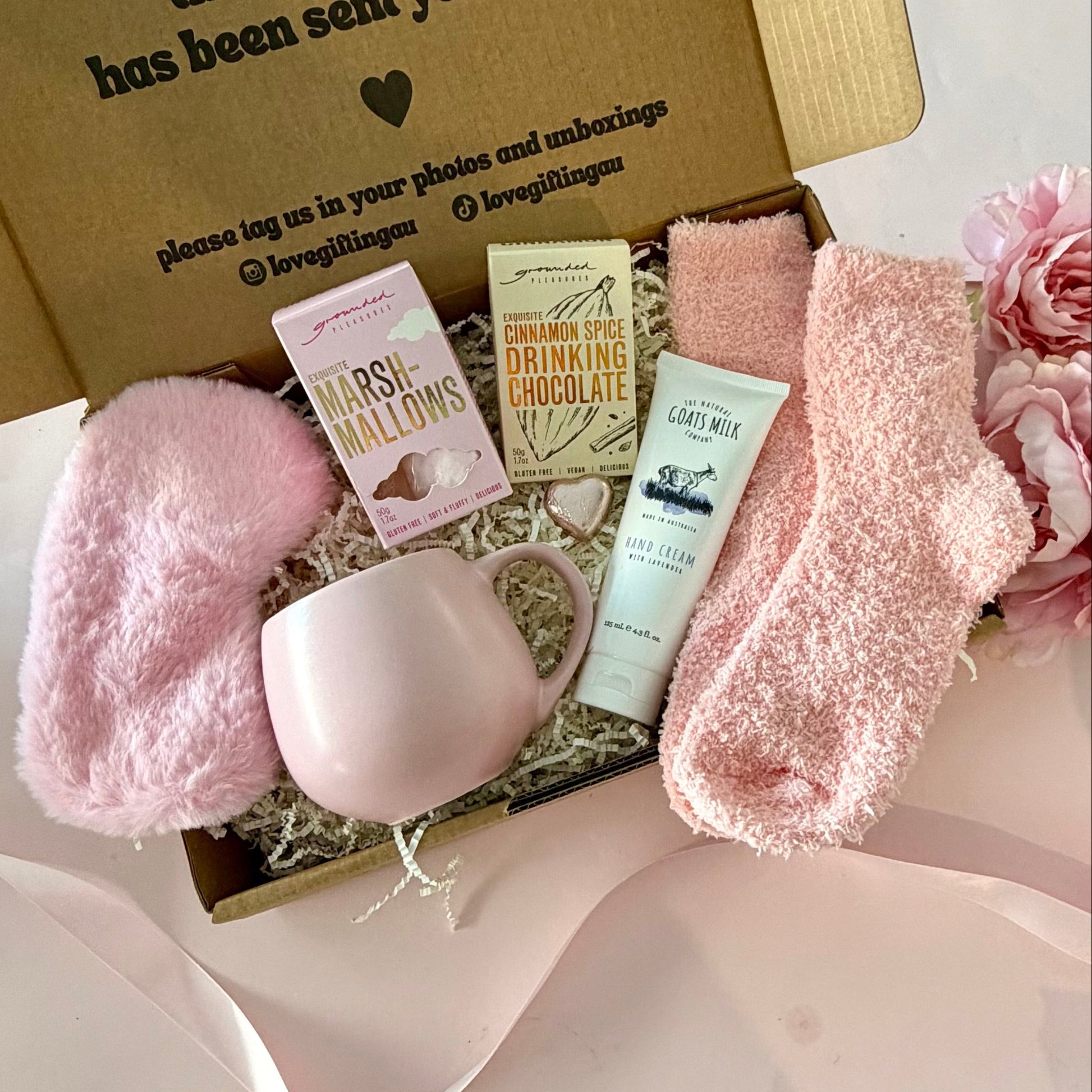 A curated comfort winter gift box featuring a pink mug, fluffy pink socks, gourmet marshmallow box, Grounded Pleasures hot chocolate box, mini chocolate heart, pink fluffy sleep mask and a lavender tube of hand cream 