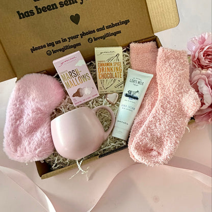 A curated comfort winter gift box featuring a pink mug, fluffy pink socks, gourmet marshmallow box, Grounded Pleasures hot chocolate box, mini chocolate heart, pink fluffy sleep mask and a lavender tube of hand cream 